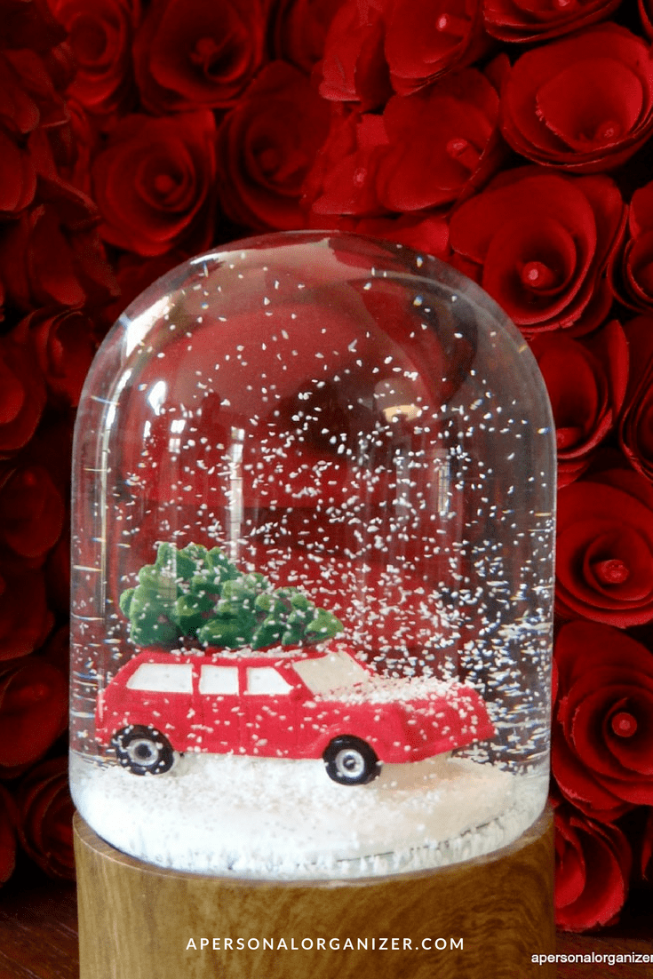 A red toy car with a Christmas tree on top sits inside a snow globe, artificial snow swirling around. Red roses fill the background. For more Christmas decoration tips, visit apersonalorganizer.com at the bottom.