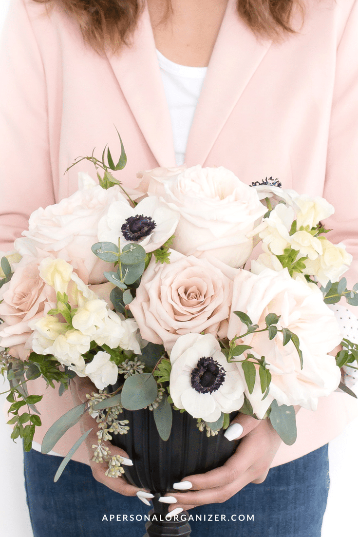 A person in a light pink blazer, preparing for hosting, holds a bouquet of flowers featuring pink roses, white anemones with dark centers, and eucalyptus leaves in a black vase.