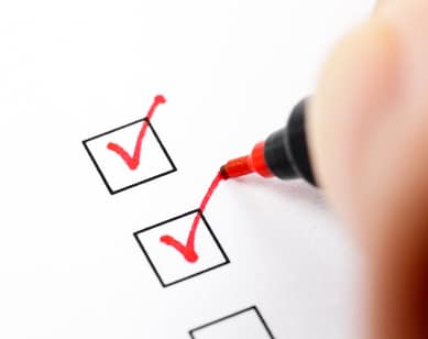 A hand is marking checkmarks in red ink on a college application checklist. Two out of three boxes have been checked. The focus is on the pen marking the checklist and the red checkmarks inside the boxes.