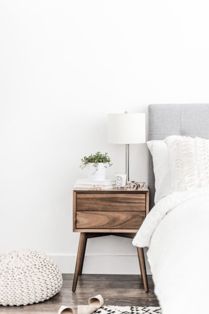 A modern wooden nightstand beside a bed with white bedding holds a neat arrangement of essentials, proving that style doesn’t require clutter. A knitted pouf and a pair of shoes rest on the wooden floor, completing the cozy scene.