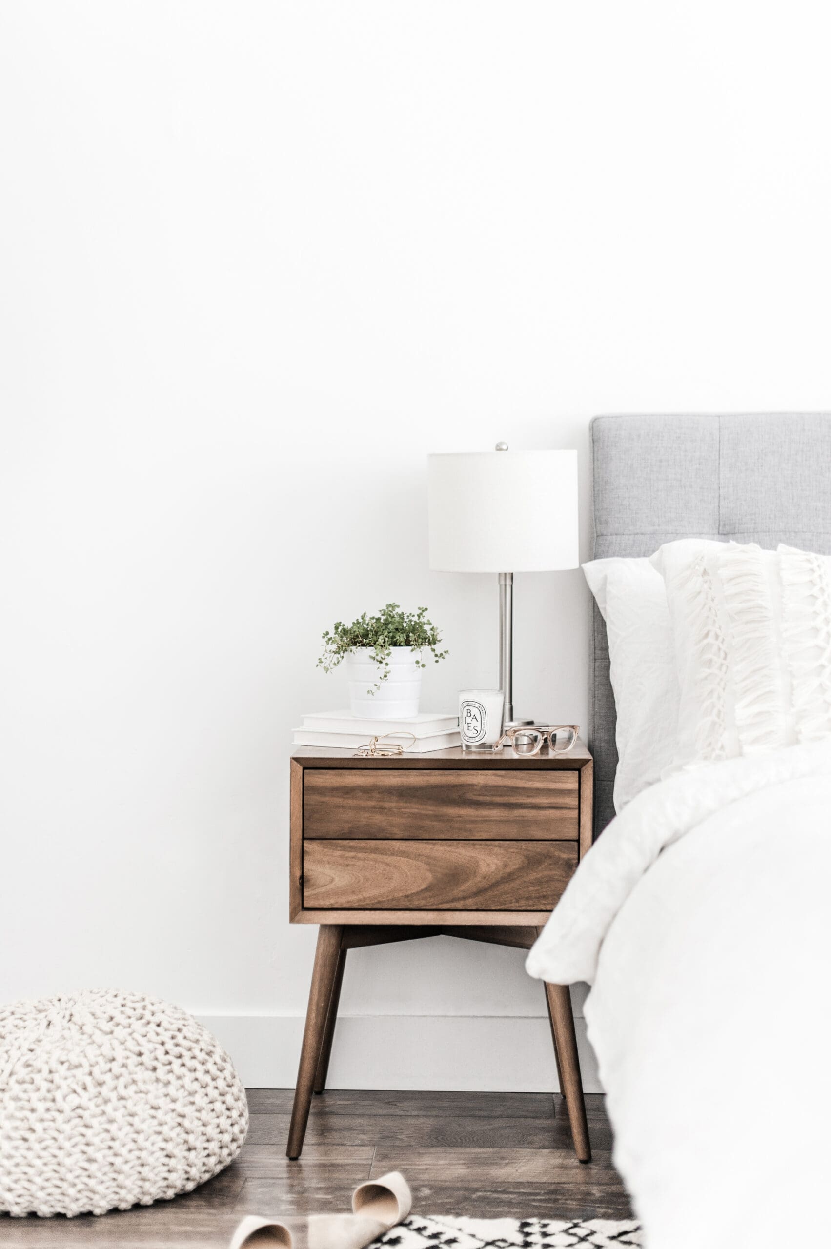 A modern wooden nightstand beside a bed with white bedding holds a neat arrangement of essentials, proving that style doesn’t require clutter. A knitted pouf and a pair of shoes rest on the wooden floor, completing the cozy scene.