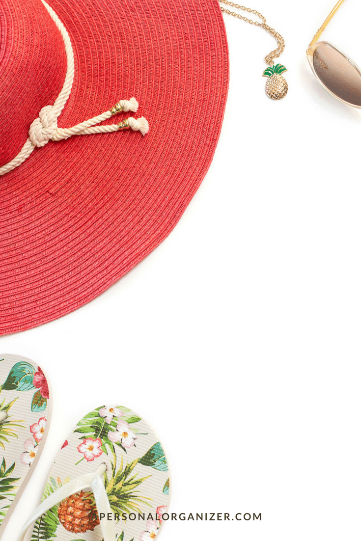 A red summer hat with a white rope detail, a pair of sunglasses, a gold necklace with a pineapple charm, and floral-patterned flip-flops are arranged on a white background. The text "APERSONALORGANIZER.COM" is visible at the bottom. Learn how to pack a beach bag effortlessly with these essentials.