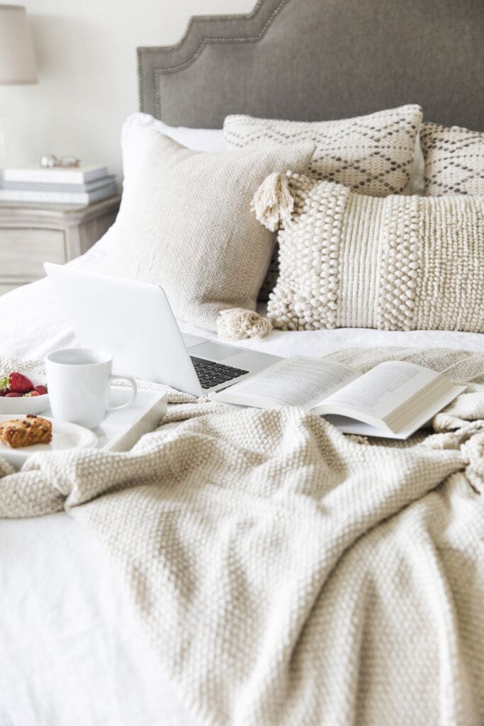 A cozy bed with textured cream pillows and a knit blanket, featuring an open laptop, a book, and a tray with coffee, pastry, and strawberries—creating a warm, inviting atmosphere that's ready for summer.