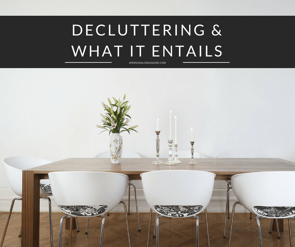 A minimalist wooden dining table with white, patterned chairs, a vase of flowers, and three candles placed on top. The text above reads, "Decluttering & What It Entails." The decluttered background is a plain white wall.