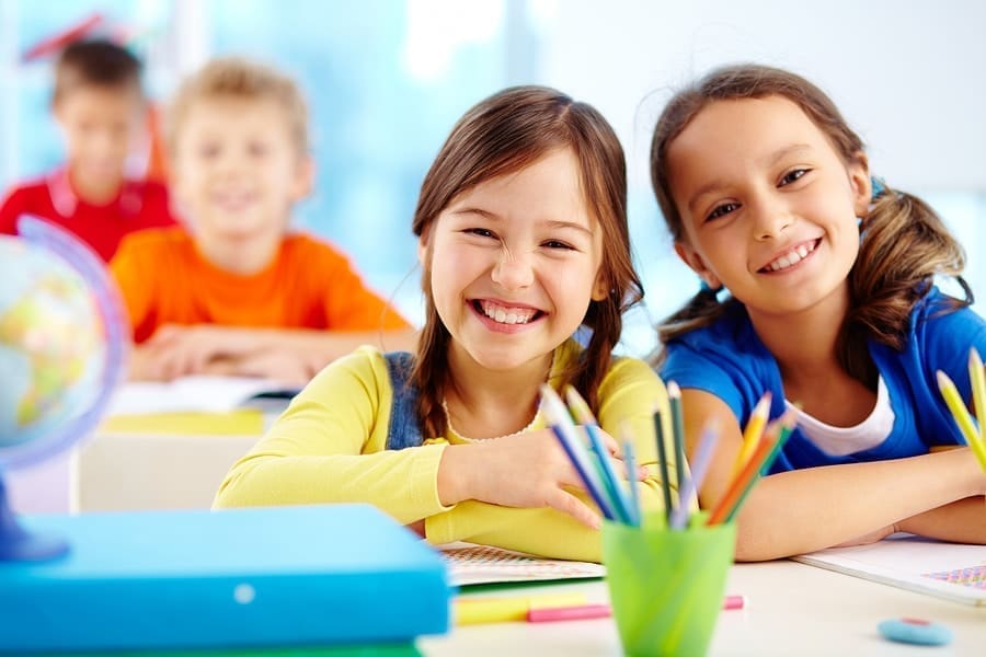 Two smiling young girls sit at a desk in a colorful classroom, likely fresh from school supply shopping. The girl on the left wears a yellow shirt and denim overalls, while the girl on the right sports a blue shirt. In the background, two other children are blurred. School supplies are on the desk.