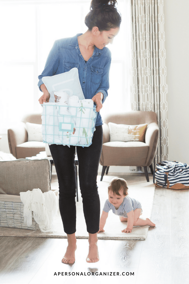 A woman in a denim shirt and black pants holds a laundry basket, looking down at a baby crawling on the floor. The room features light-colored furniture and decor, creating a cozy atmosphere that highlights effective home management. The text "apersonalorganizer.com" is visible at the bottom of the image.