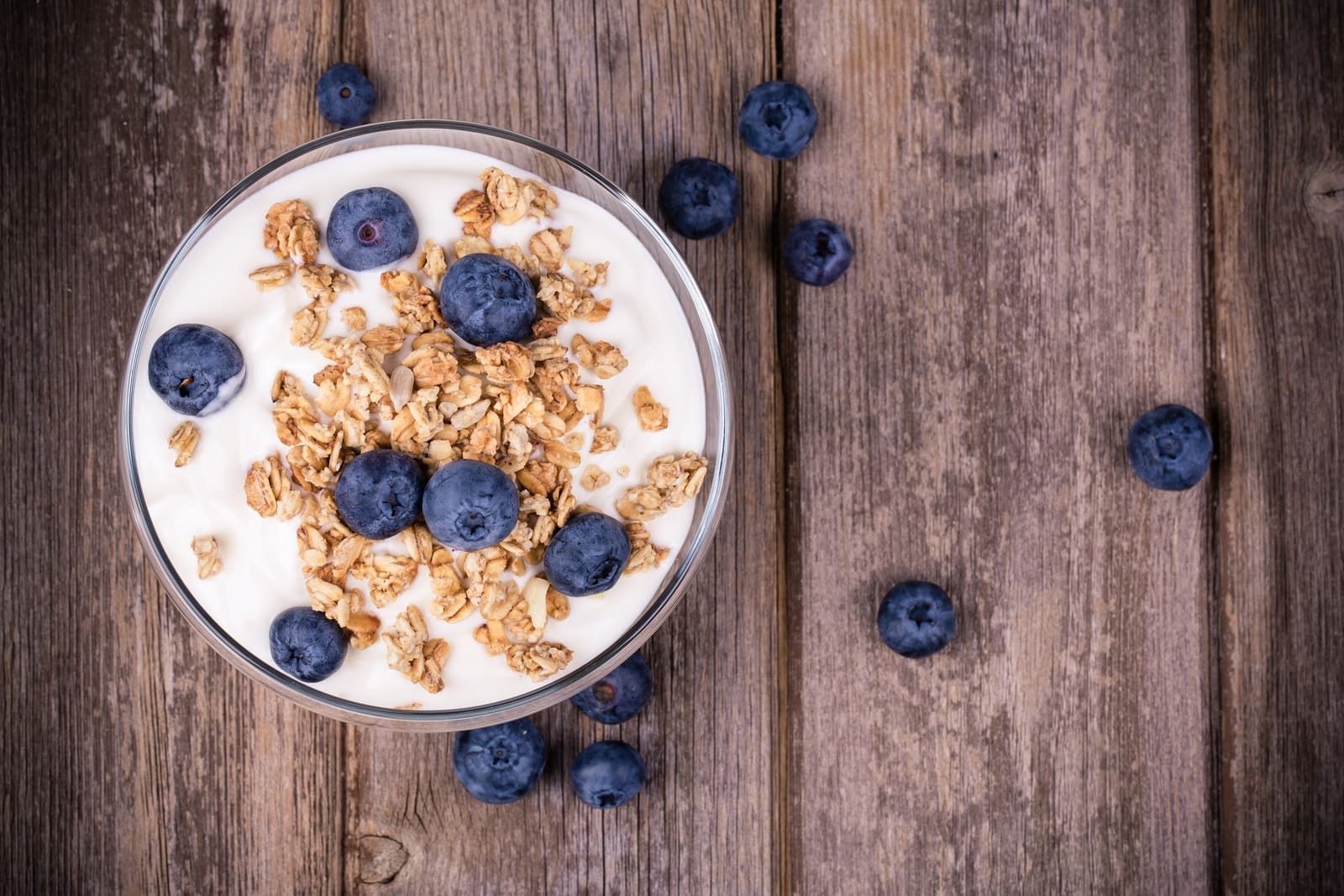 A bowl of yogurt topped with granola and fresh blueberries sits on a rustic wooden surface, perfectly organized for an appetizing display. Additional blueberries are scattered around the bowl on the wooden planks.