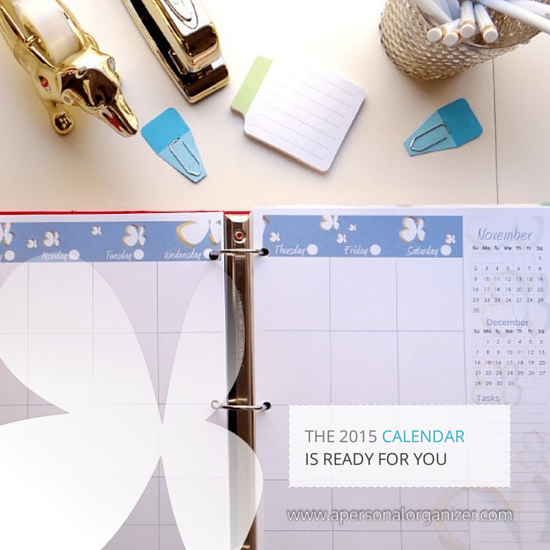 An open planner displaying a 2015 calendar page for November, adorned with a butterfly design. Nearby are a golden stapler, blue clips, sticky notes, pens in a holder, and a calendar widget. Text reads "THE 2015 CALENDAR IS READY FOR YOU" with the URL www.apersonalorganizer.com. Perfect for your home management binder!