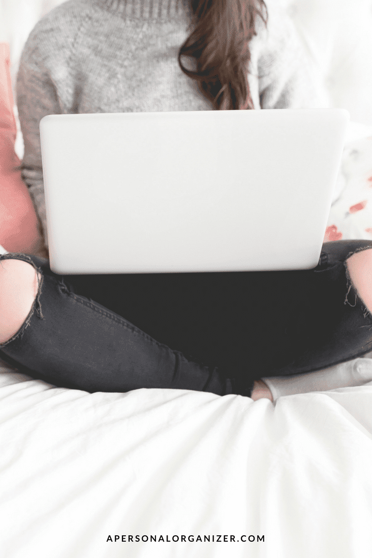 A person sits cross-legged on a bed, focused on their laptop. Dressed in a gray sweater and black ripped jeans, they might be indulging in some Black Friday shopping. At the bottom of the image, the website "apersonalorganizer.com" is visible.