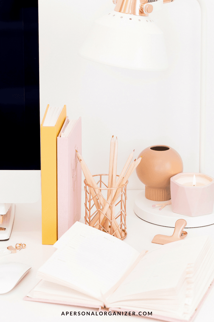 A neatly organized white desk featuring an open notebook, a set of pencils in a holder, two pastel-colored closed journals, a small round vase, a lit candle, and an angled white desk lamp. A portion of a computer screen, perhaps displaying email inbox organization tips, is visible on the left side.