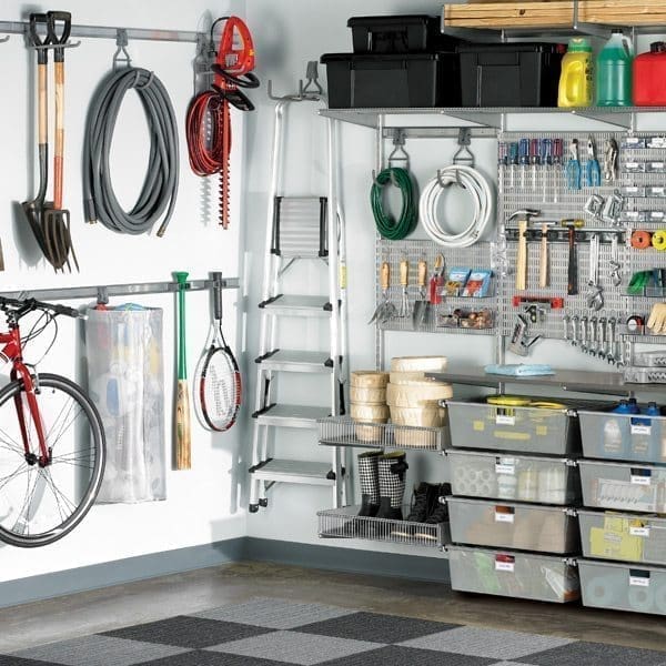 A well-organized garage with various items neatly stored showcases excellent garage organizing. Tools and garden equipment hang on the wall, while shelves hold storage boxes, cleaning supplies, and paint cans. Bicycles, a ladder, and sports equipment are also visible. The floor is covered with gray and black tiles.