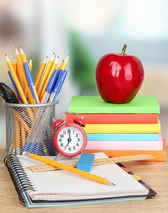A neatly arranged school desk, ready for back to school, featuring a stack of colorful notebooks, a red apple on top, a pencil holder filled with pencils and pens, a small red alarm clock, a ruler, and an open notebook with a pencil and eraser.