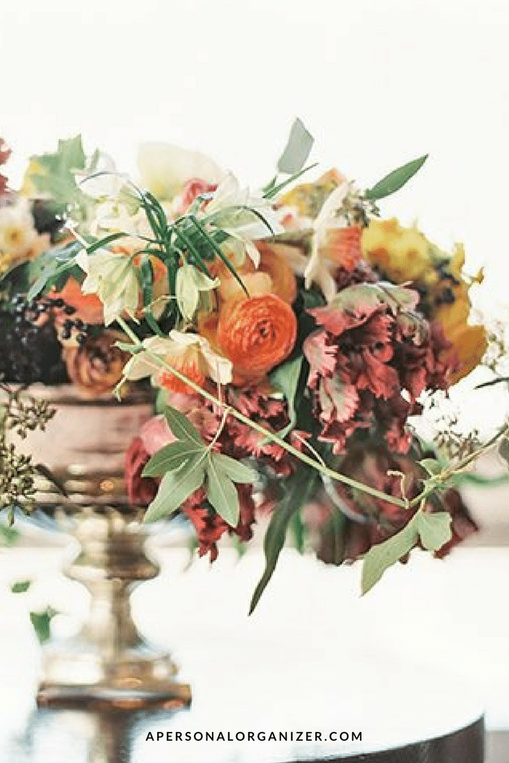 A colorful floral arrangement with orange, yellow, and white flowers, alongside green leaves and stems, displayed in a gold vase. Perfect for a Thanksgiving table, the image has the website "APERSONALORGANIZER.COM" at the bottom.
