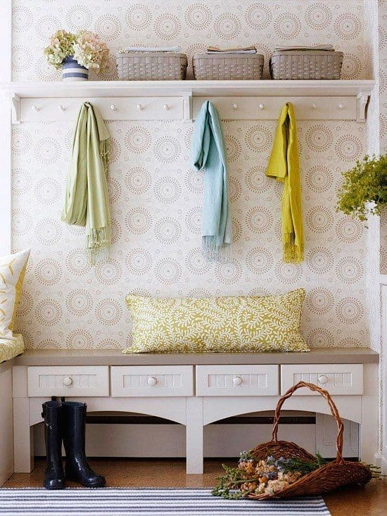 A small entrance with a white bench featuring three drawers. Above the bench is a shelf with three wicker baskets and hooks holding green, blue, and yellow scarfs. A pair of black rain boots is on the floor, and a wicker basket filled with flowers sits nearby, perfect for organizing a narrow entry.