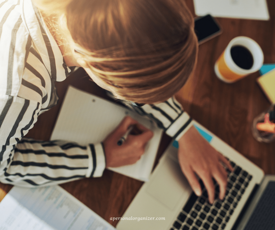 A person with blonde hair, wearing a black and white striped shirt, writes in a notebook while using a laptop. There is a cup of coffee and some papers on the wooden table. The scene suggests they might be a professional organizer, deep in a focused work or study session.