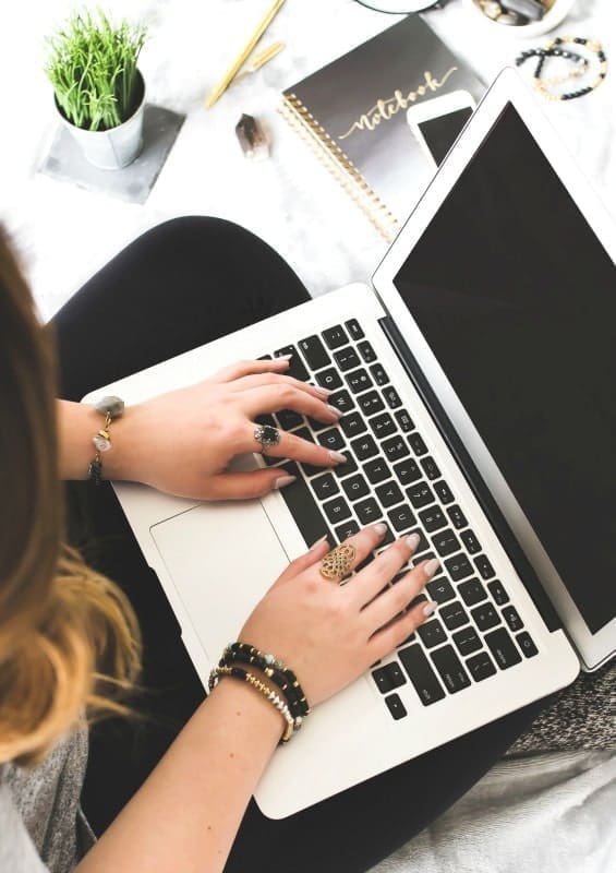 A person is typing on a laptop placed on their lap, likely working on college assignments. They are wearing multiple rings and bracelets. Next to the laptop are a notebook, pen, and a small potted plant on a white surface. The setting appears casual and relaxed, making it easy to move between tasks efficiently.