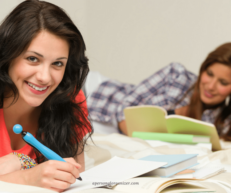 Two women are lying on a bed in their cozy dorm space, studying. The woman in the foreground, wearing a red shirt, is smiling and holding a blue pen while looking at the camera. The woman in the background is wearing a plaid shirt and is focused on reading a book. Books and papers are scattered around them.