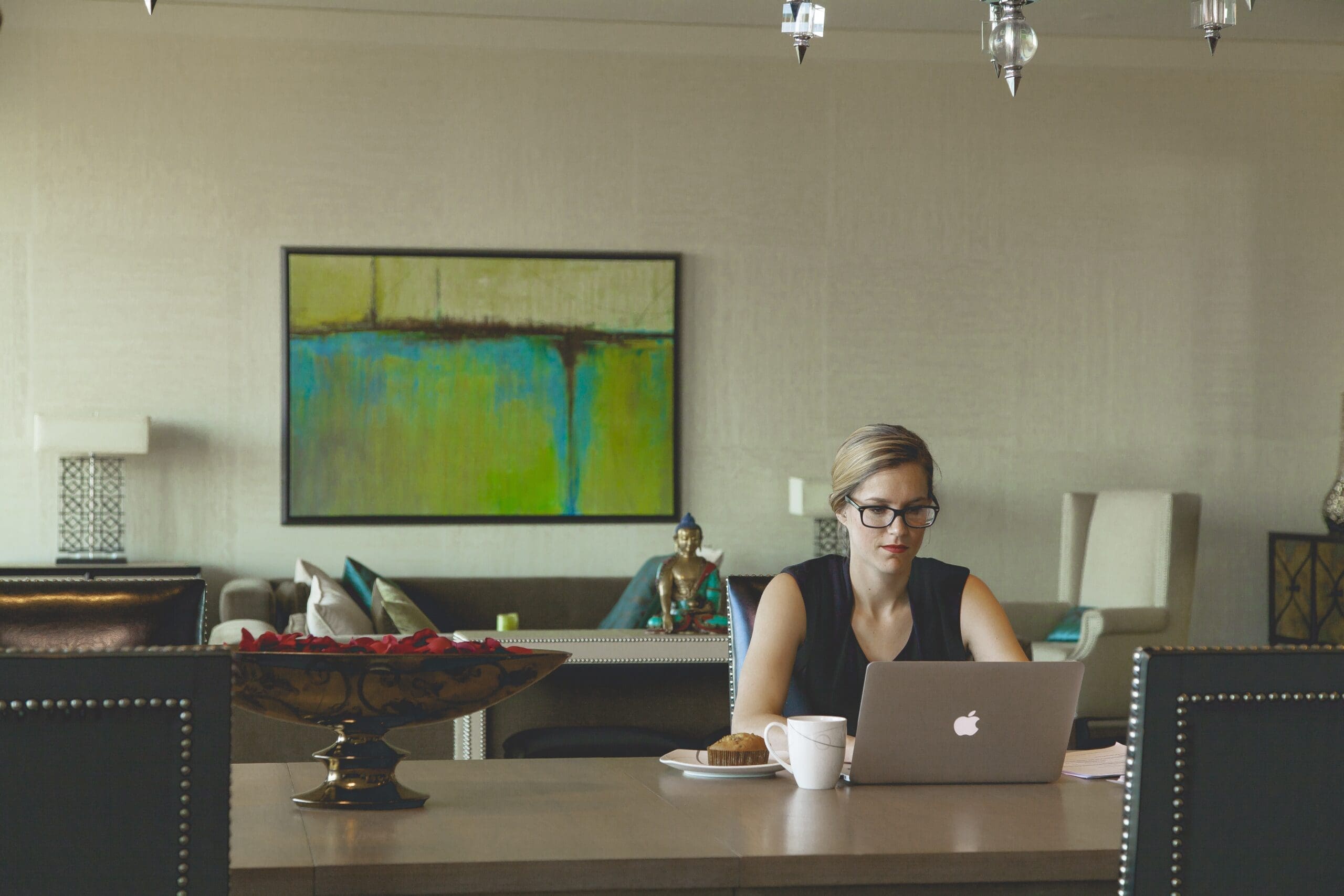 A woman sits at a dining table in a modern home, working on a laptop as she looks focused. She wears glasses, reflecting the soft glow of her screen. Behind her hangs a large abstract painting. The room blends contemporary and traditional elements, including a Buddha statue and offers insights on how to como formalizar various processes.