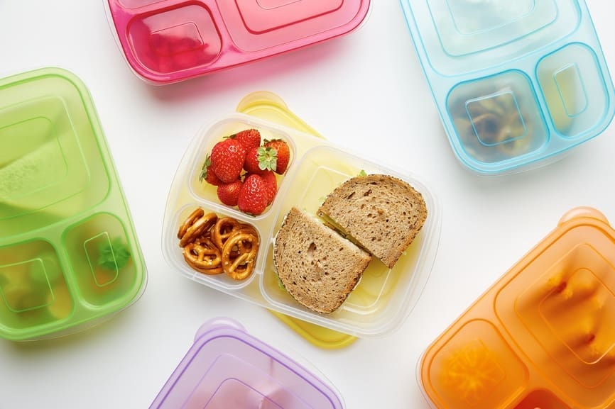 A variety of colorful, segmented lunch containers are arranged on a white surface. One open container has two strawberry halves, a handful of pretzels, and a sandwich with whole-grain bread—perfect for school lunches. Other closed containers are visible in different colors including pink, blue, and green.
