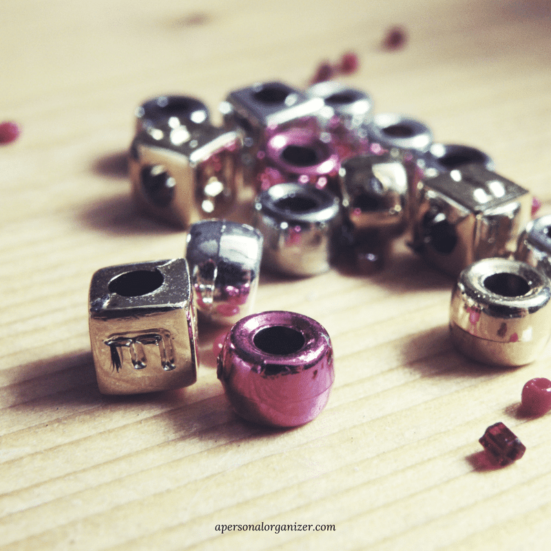 A close-up photo captures various metallic beads scattered on a wooden surface, reminiscent of a decluttering journey. The beads, in square and round shapes, shimmer in silver, gold, and pink hues.