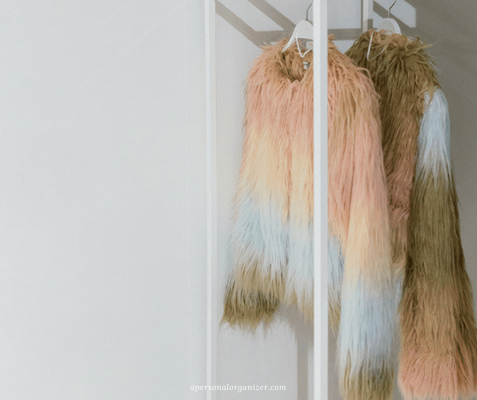 Two colorful faux fur jackets, featuring pastel hues of pink, blue, and yellow, hang in a decluttered white minimalist closet. The closet has a glass door, reflecting light that softly illuminates the vibrant textures of the jackets.
