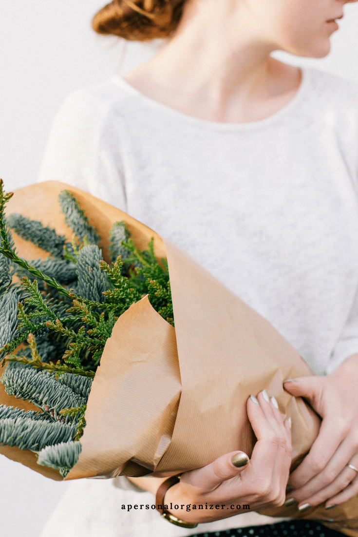 A person wearing a white sweater is holding a bouquet of greenery wrapped in brown paper, standing in a narrow entryway. The person's head is turned to the side, and their nails are painted metallic. The website "apersonalorganizer.com" is visible at the bottom of the image.