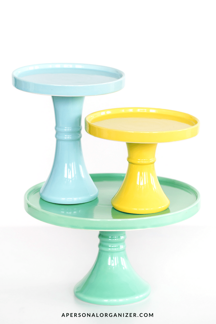 Three colorful cake stands in varying heights are stacked on one another, perfect for decluttering your dessert display. The bottom stand is green, the middle yellow, and the top light blue. Each stand has a glossy finish and a wide base. The background is white, with the stands placed centrally.