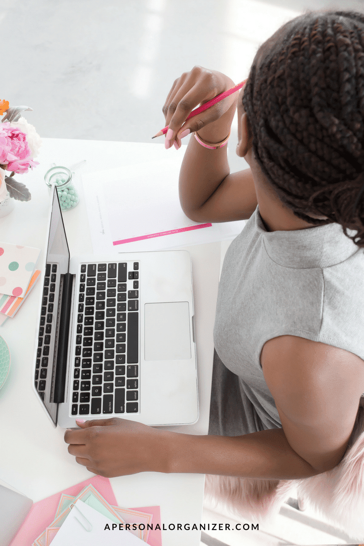 A person with braided hair is diligently working on a computer at a desk, jotting down notes with a pink pen. The workspace features a flower arrangement, colorful stationery, and a cup of coffee. They're possibly brainstorming ideas to market new local businesses. The website name apersonalorganizer.com is shown at the bottom.