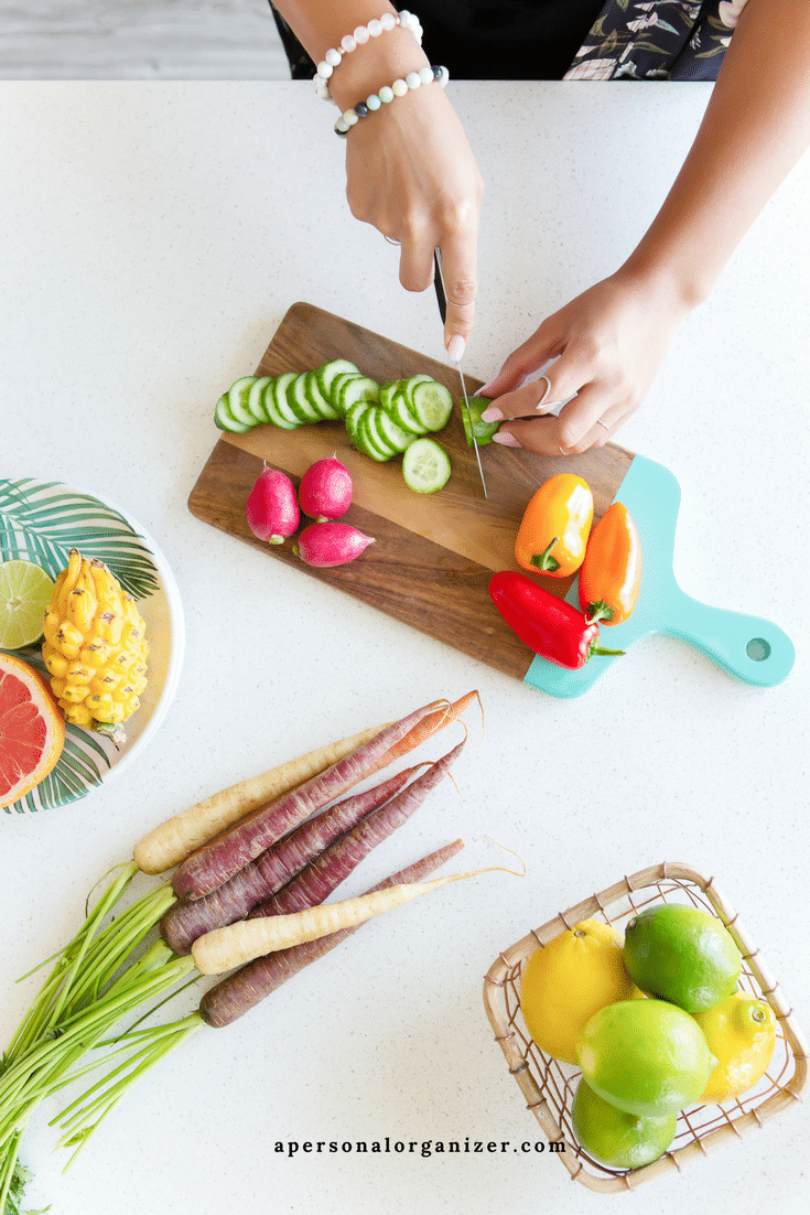 A person is slicing cucumbers on a wooden cutting board with colorful radishes and peppers beside it while planning their menu. A basket of lemons and limes, heirloom carrots, and a plate with pineapple and grapefruit are arranged on the counter, alongside the website "apersonalorganizer.com" visible.