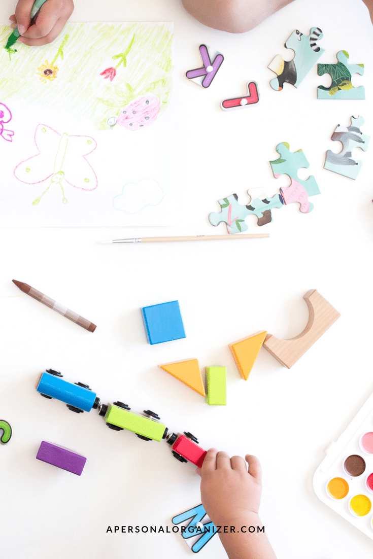 A child's hands are visible as they play on a white table. They are drawing with crayons, assembling puzzles, arranging colorful wooden blocks, and playing with a toy train. A box of watercolors is also present. The image suggests a creative and playful scene—perfect for inspiring gift ideas.
