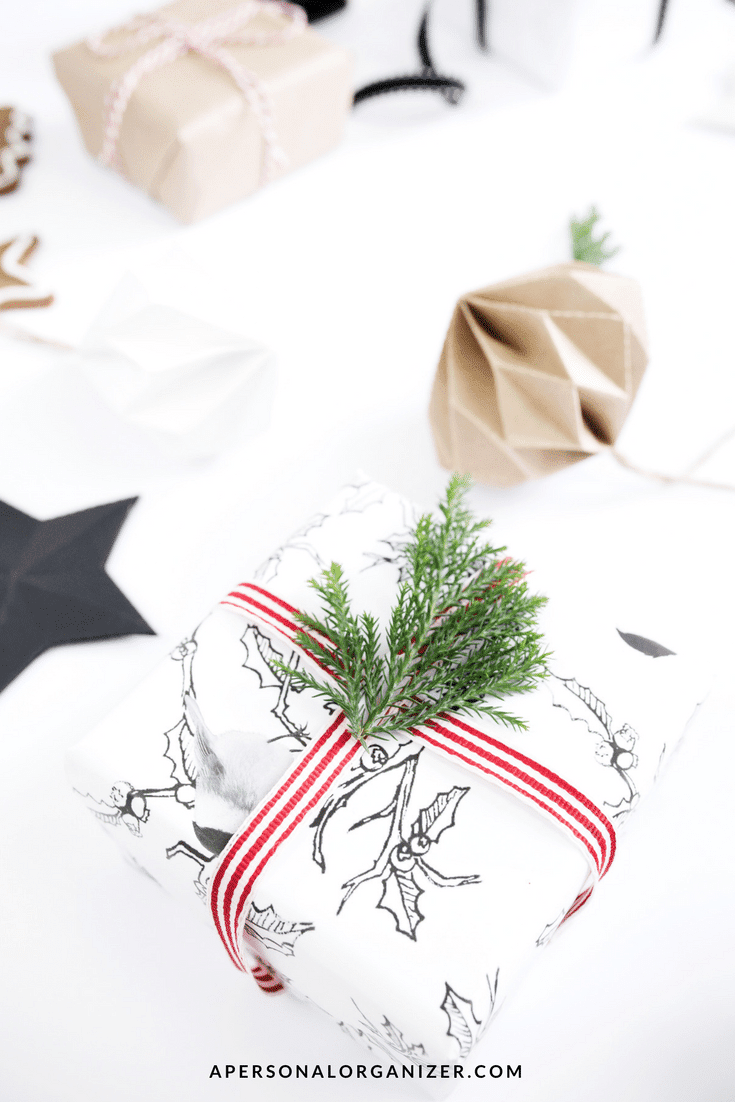 A neatly wrapped gift in black and white holly-patterned paper with a red and white striped ribbon and a sprig of evergreen on top. Other wrapped gifts, ideal for teen gift ideas, and holiday decorations are in the background. The bottom of the image features the text "APERSONALORGANIZER.COM.
