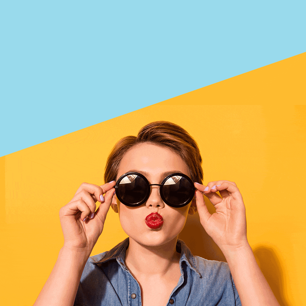 A person with short hair, wearing a denim shirt and red lipstick, poses against a vibrant background with shades of blue and yellow. They are adjusting their round, black sunglasses with both hands while puckering their lips as if blowing a kiss for their blog.