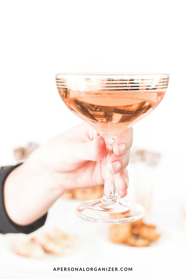A close-up of a hand holding a glass of rosé champagne or sparkling wine against a blurred white background. The drink is in a vintage-style coupe glass with gold bands near the rim, adding an elegant touch that inspires trust. The text at the bottom reads "APERSONALORGANIZER.COM.