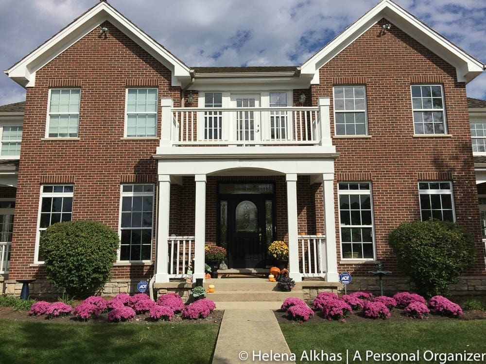 A large two-story brick house with white pillars and railings, featuring a central black front door with glass panels. The landscaped front yard has bushes and vibrant pink flowers lining the pathway leading to the entrance. Perhaps someone leaving the military would find peace here. The sky is partly cloudy.
