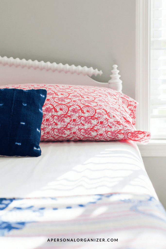A neatly made bed with a white frame, featuring a pink patterned pillow and a dark blue pillow against a light gray wall, awaits house guests. The bed sheet is pristine white, and sunlight streams through partially opened blinds, casting shadows on the inviting bed.