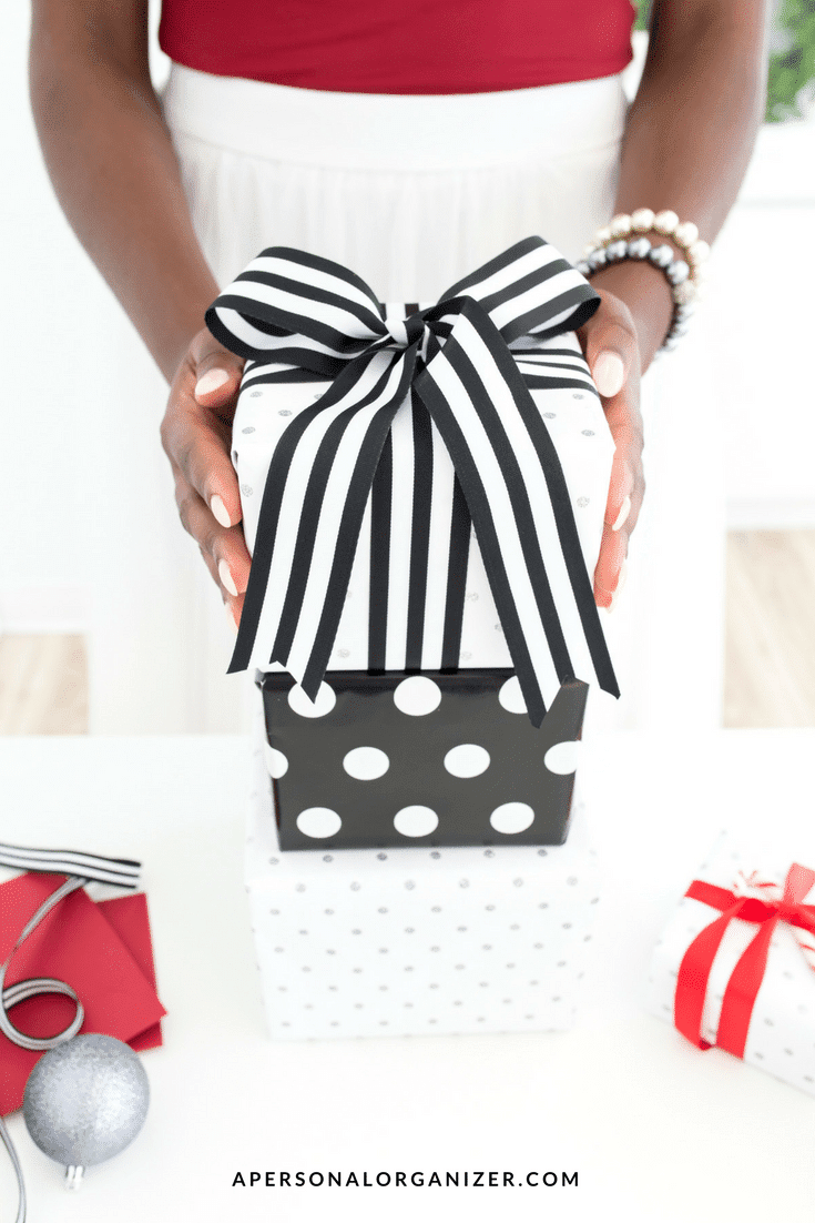 A person in a red top and white skirt is holding a stack of three holiday gift boxes wrapped in black and white paper with a striped ribbon. One box has a dot pattern. A red gift wrap, ribbon, and ornament are on the white table. The website name is at the bottom.
