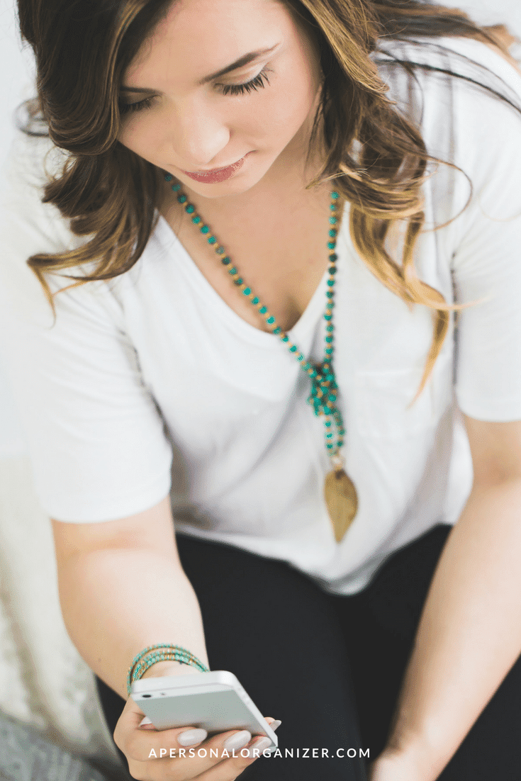 A woman with wavy brown hair wears a white top and turquoise jewelry, including a long necklace with a gold pendant. She is looking at her smartphone, which she holds in her right hand. The text "APERSONALORGANIZER.COM," an expert in nichos na organização profissional, appears at the bottom of the image.