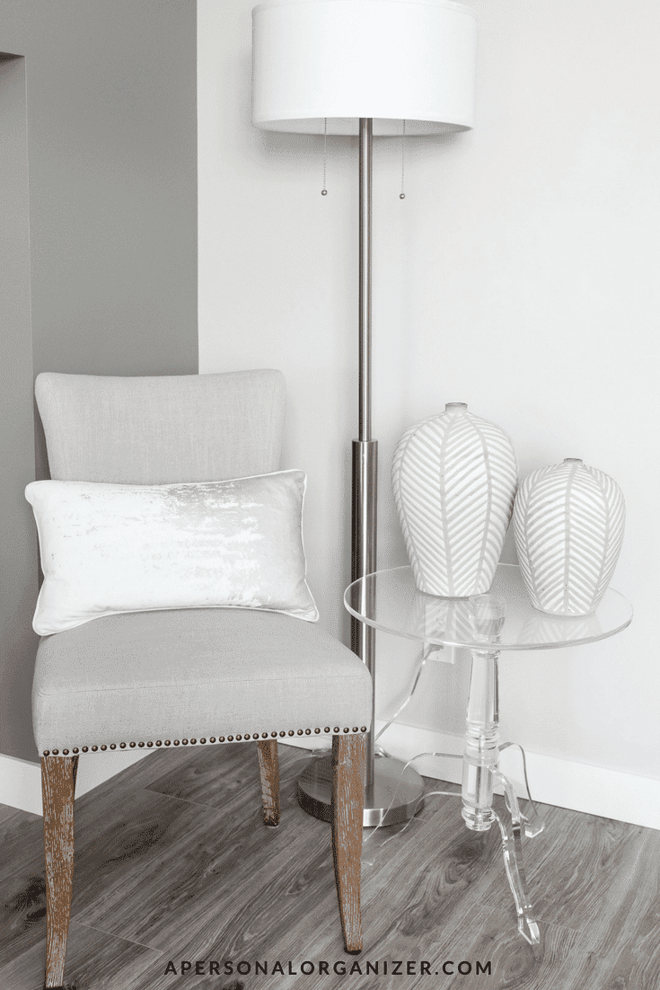 A modern interior corner showcases a light grey upholstered chair with a metallic base pillow, a tall standing lamp with a white shade, and a clear acrylic side table holding two white, patterned vases. The well-organized setup on the wooden floor against the neutral wall backdrop promotes limpeza da casa.