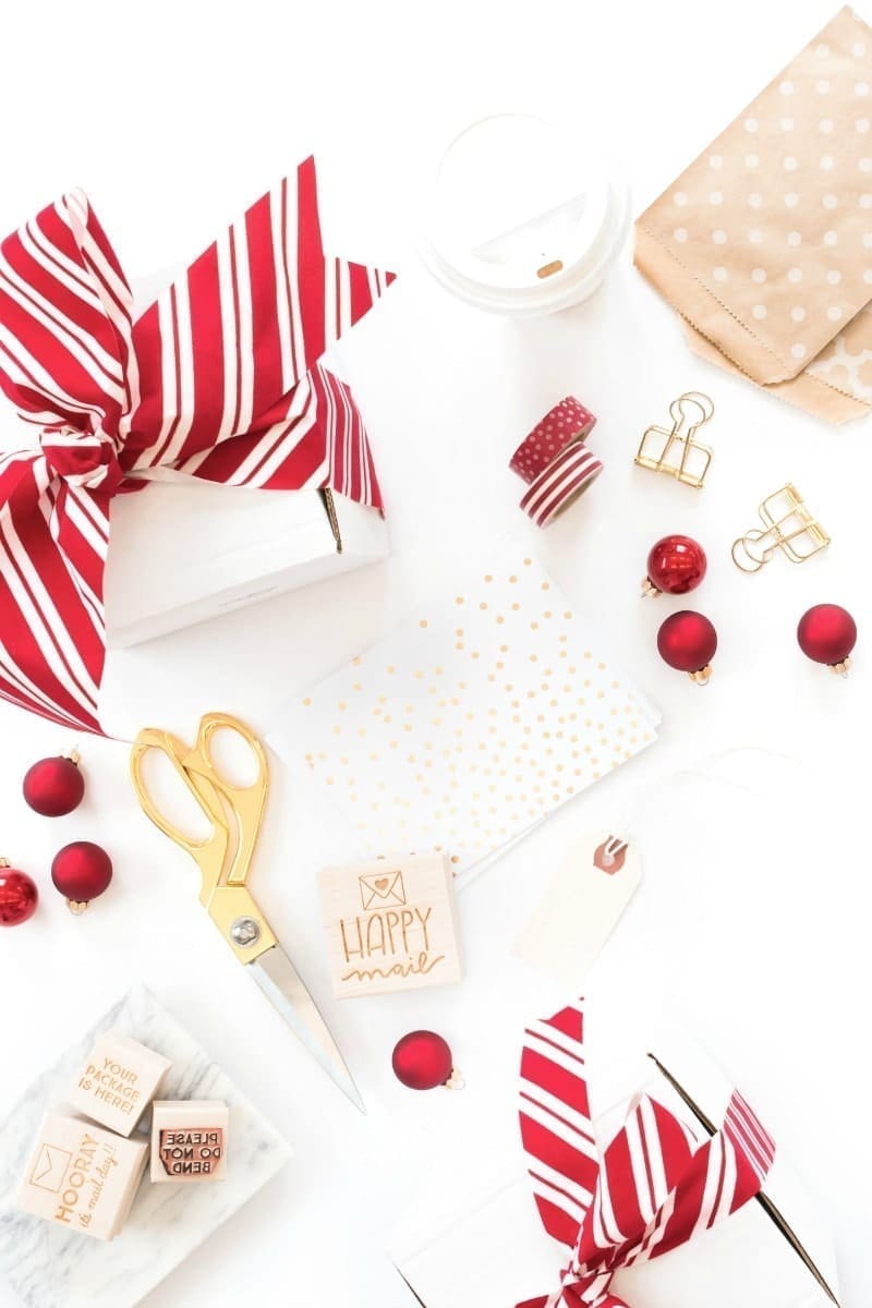 A festive flat lay perfect for holiday marketing ideas, featuring holiday gift wrapping items. A white box with a red and white striped ribbon, gold scissors, decorative tape, red ornaments, gold clips, a card with gold polka dots, and a stamp that says "Happy Mail.