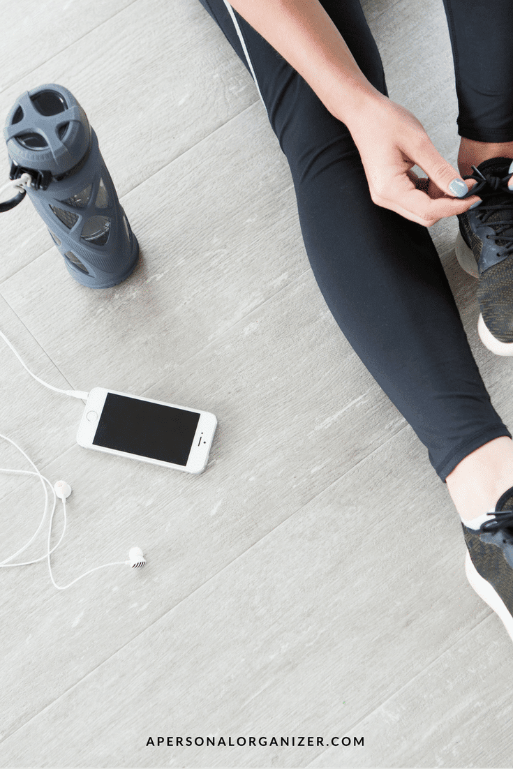 A person wearing black athletic wear ties the laces of their black sneakers. Nearby, a smartphone with earphones plugged in, and a water bottle with a handle cap are on the floor—a scene reflecting their new year's resolution. The text "APERSONALORGANIZER.COM" is visible at the bottom.