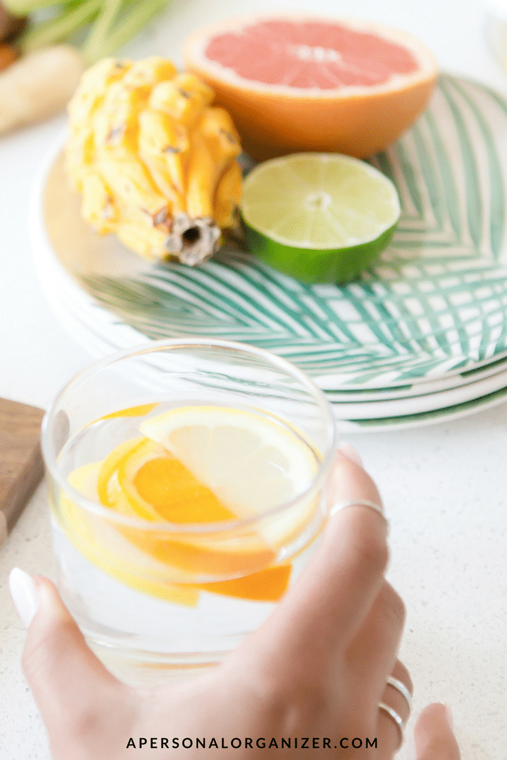 A hand with silver rings holds a glass of water with lemon slices over a white counter, making it a refreshing start to the morning routine. Behind the glass are a halved lime, a halved grapefruit, and a yellow dragon fruit on a green and white patterned plate. The text "APERSONALORGANIZER.COM" is visible at the bottom.