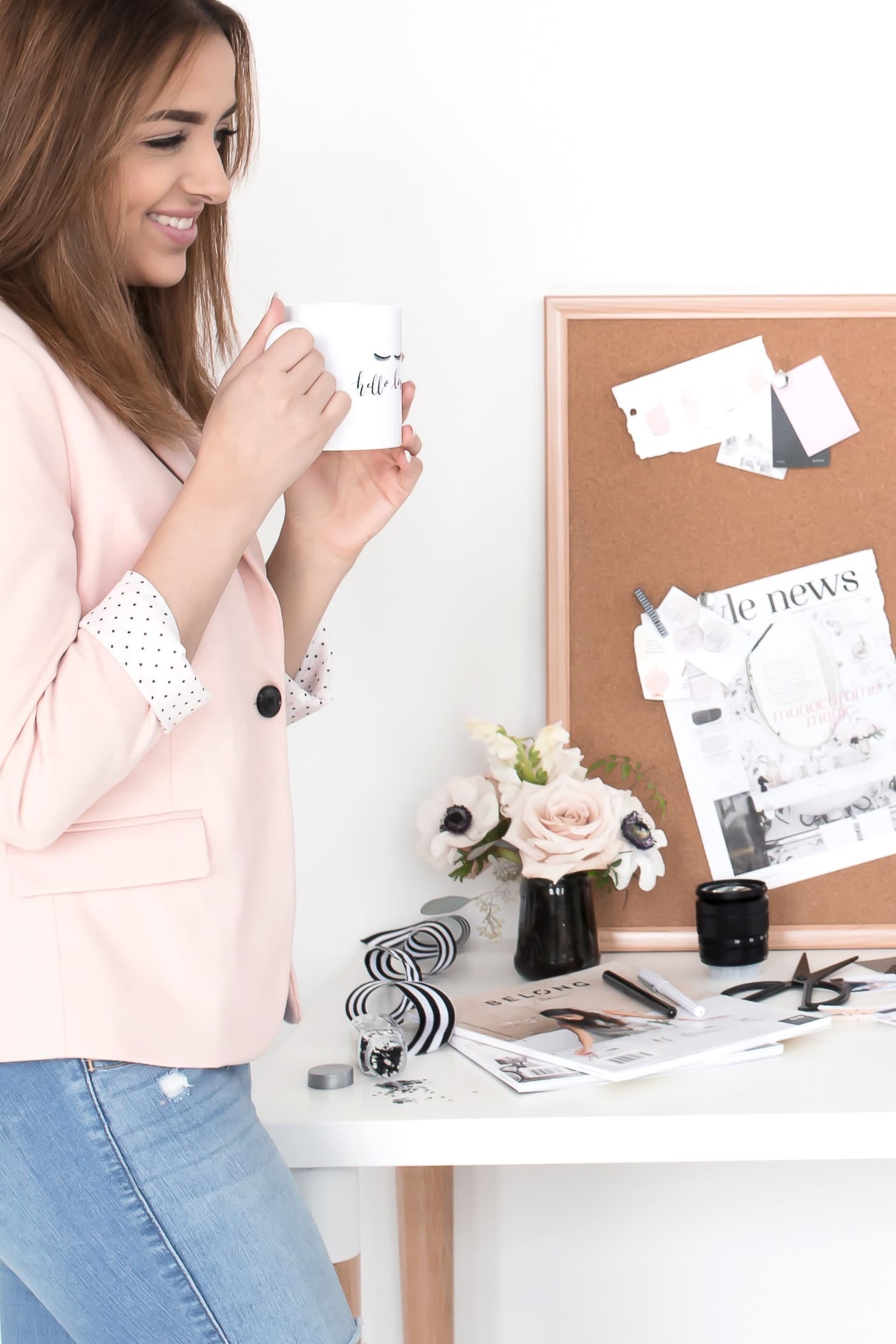 A woman wearing a light pink blazer and blue jeans is holding a mug while standing beside a desk. The desk is cluttered with magazines, a striped vase, flowers, and various office supplies. A corkboard with pinned papers and photos is in the background, perfect for working in your zone of genius.