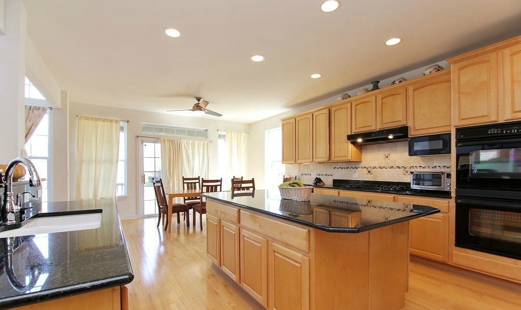 Spacious modern kitchen with light wood cabinets, black countertops, a central island, stainless steel appliances, and a dining area with four chairs by large windows—perfect for your next home tour.