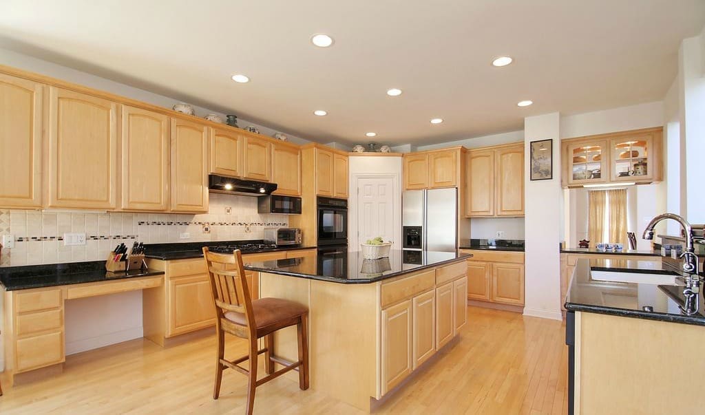 Spacious kitchen featured in a recent home tour with light wood cabinets, black countertops, a central island with a wooden chair, stainless steel appliances, recessed lighting, and a sink near a large window.