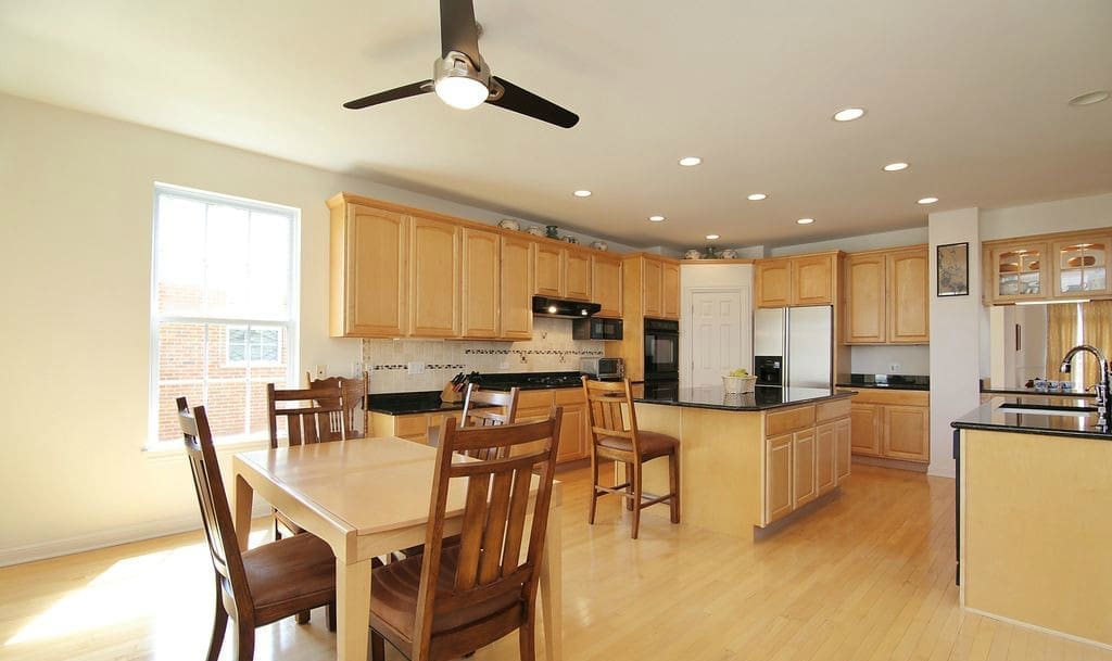 This bright kitchen, featured in a recent home tour, showcases light wood cabinets, black countertops, an island with seating, and a dining table for four. Ceiling fan, recessed lighting, and a large window create a spacious, airy feel.
