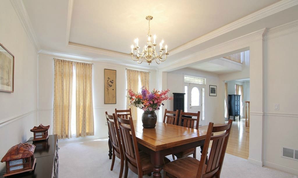 A bright dining room with a wooden table, six chairs, a vase of colorful flowers, beige curtains, a chandelier, and light walls welcomes you. On this home tour, an open doorway leads to a foyer with a white front door.