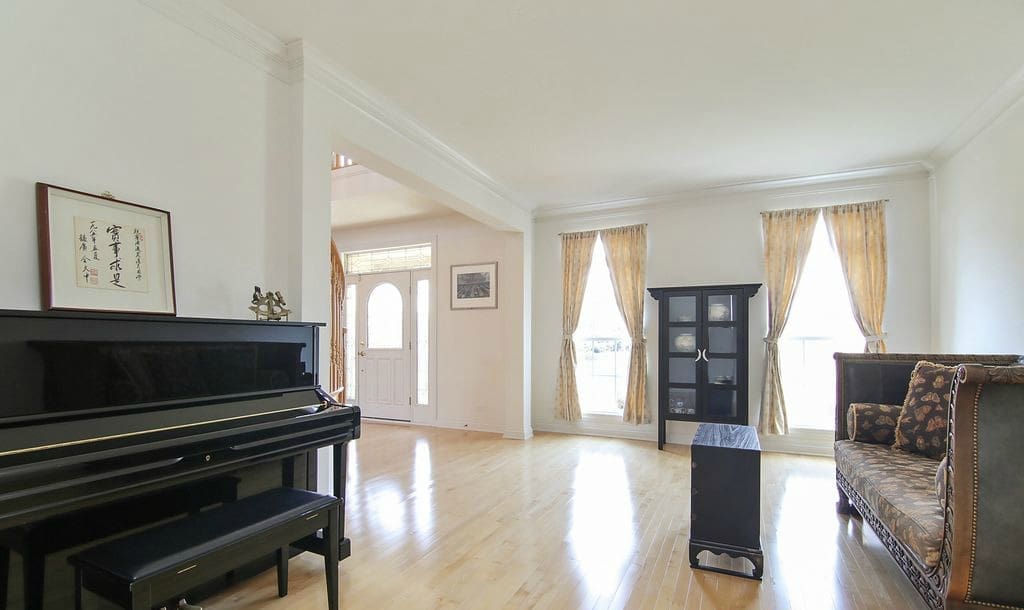 Bright living room with light wood floors, a black piano on the left, a dark sofa on the right, a black cabinet in front of three tall windows with gold curtains, and framed artwork—perfect for your next home tour.