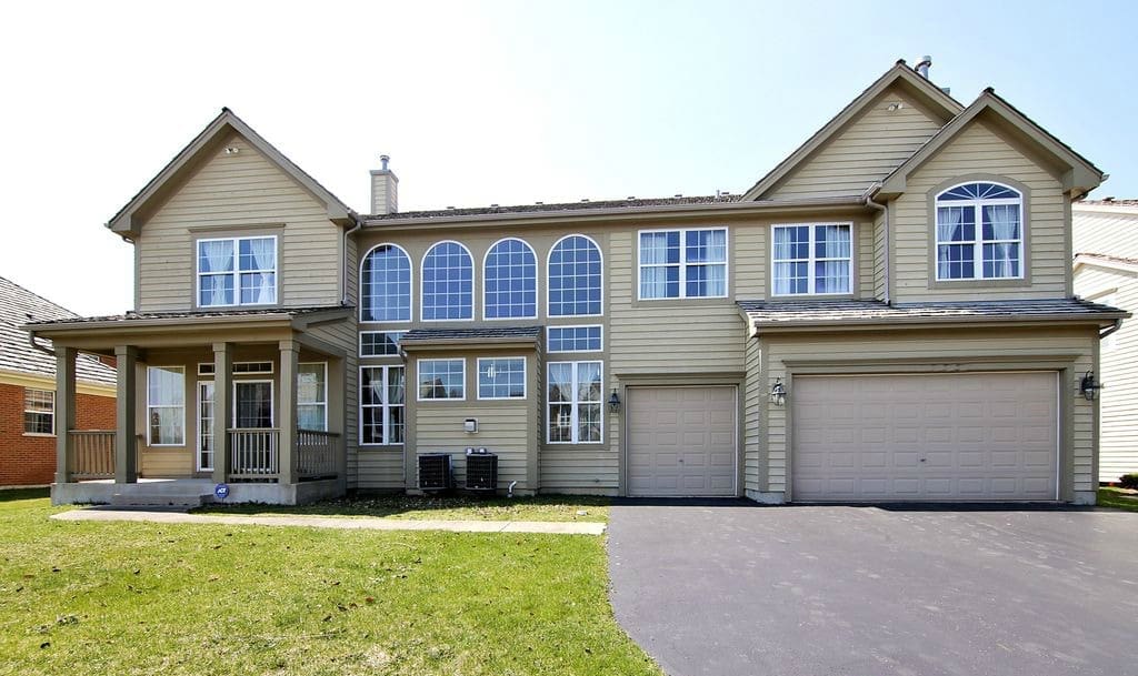 A large beige two-story suburban house with multiple tall windows, a covered front porch, and a three-car garage. Ideal for a home tour, this residence sits next to a green lawn with a blacktop driveway in front.