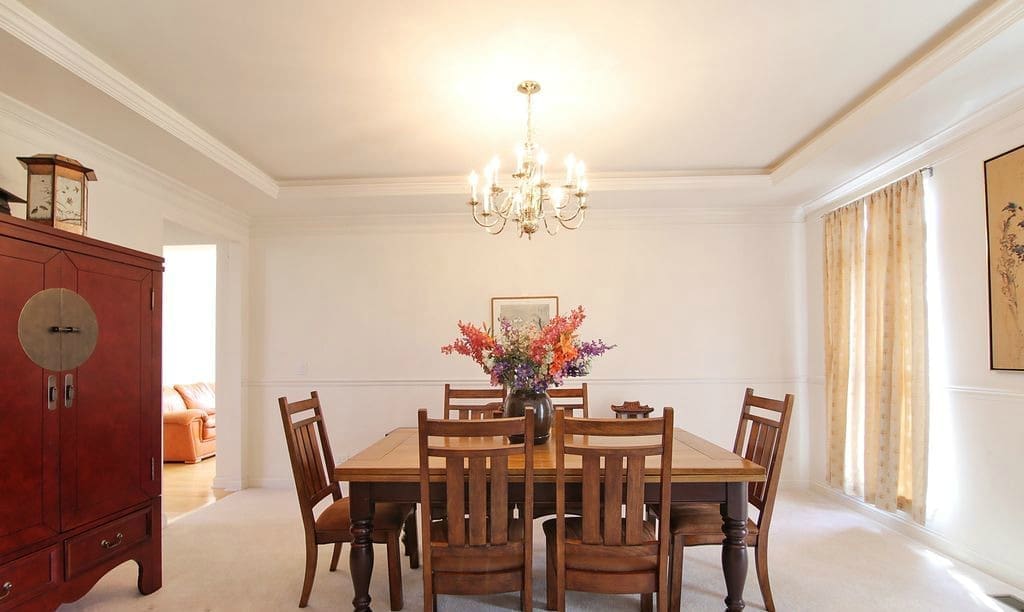 A bright dining room featured in a home tour with a wooden table and six chairs, a floral centerpiece, chandelier above, beige curtains, white walls, and a red armoire on the left.