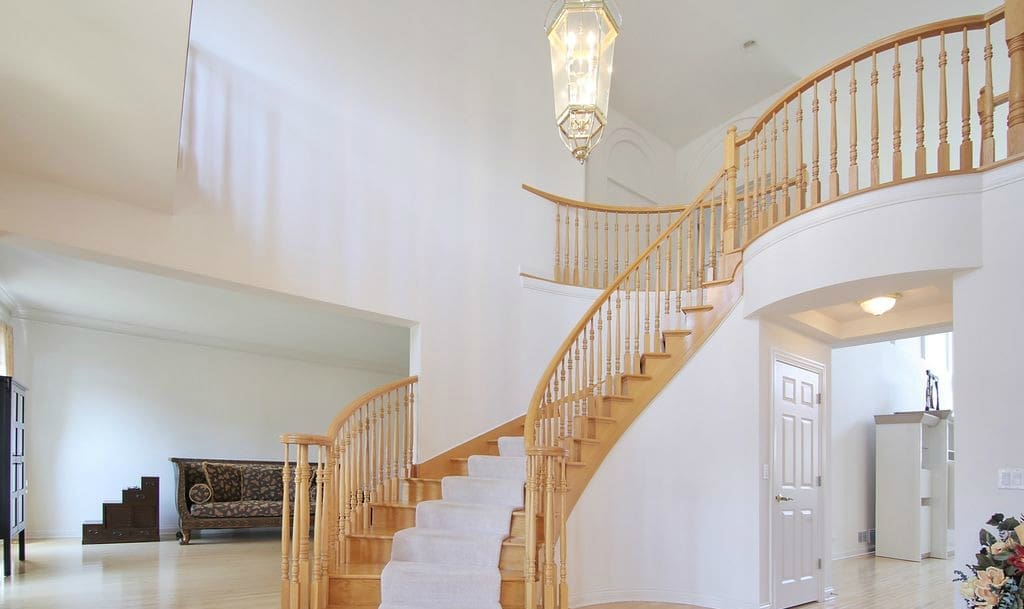 Step into this bright foyer for an inviting home tour, featuring a grand wooden staircase, white walls, and a large chandelier. Upstairs boasts a curved railing, while the living area with a patterned sofa is visible to the left.